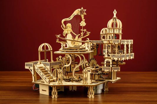 Modèle mécanique en bois de « Le jardin magique », mettant en scène une sirène harpiste entourée de structures miniatures et d’escaliers détaillés, posé sur une table devant un fond rouge.